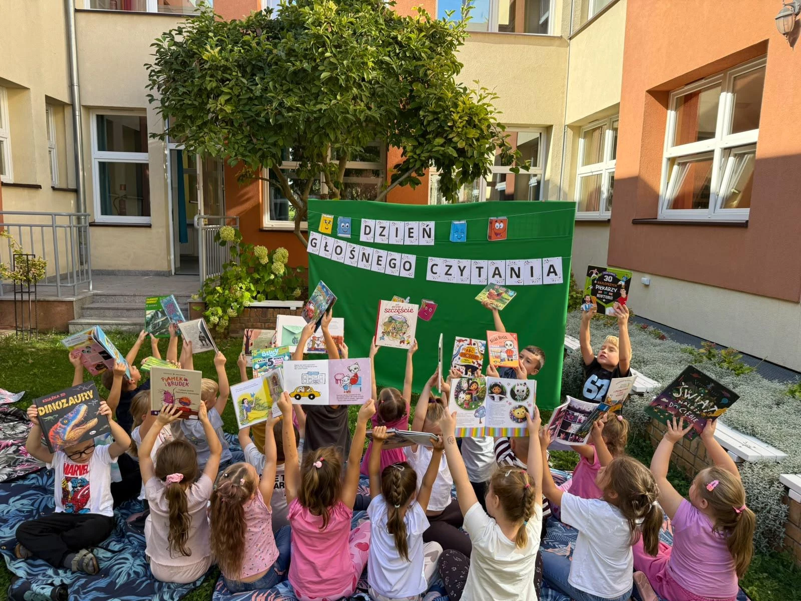 Dzieci siedzą na dworze i unoszą książki w górę.