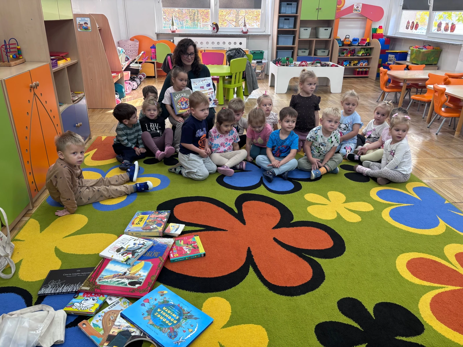 Bibliotekarka czyta książkę grupie dzieci siedzących na kolorowym dywanie.